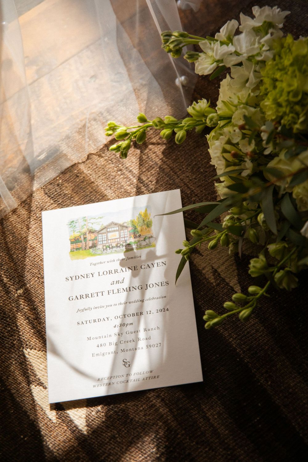 Elegant wedding invitation with watercolor venue illustration next to white flowers on burlap fabric in sunlight.