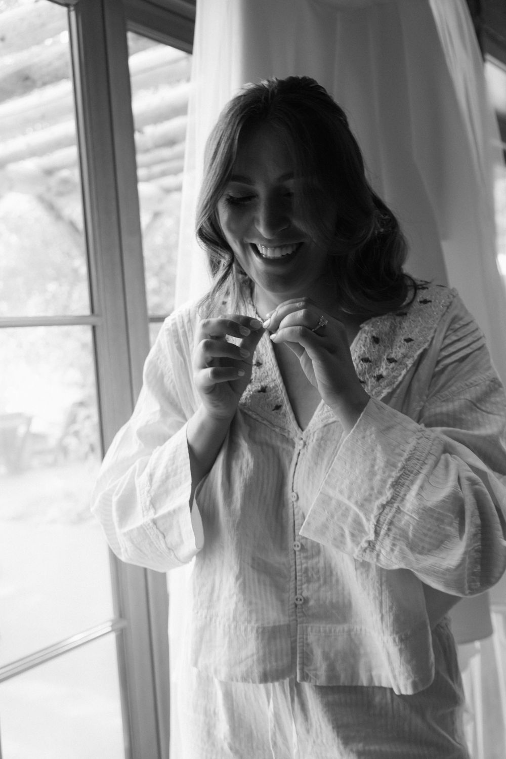 A black and white portrait of someone in white clothing smiling by a window with natural light.