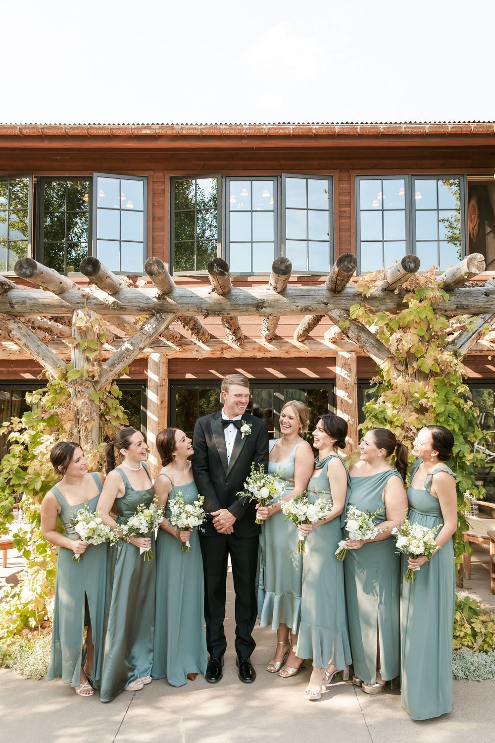 Wedding party in sage green bridesmaid dresses and black tuxedo pose under rustic wooden pergola with lush greenery.
