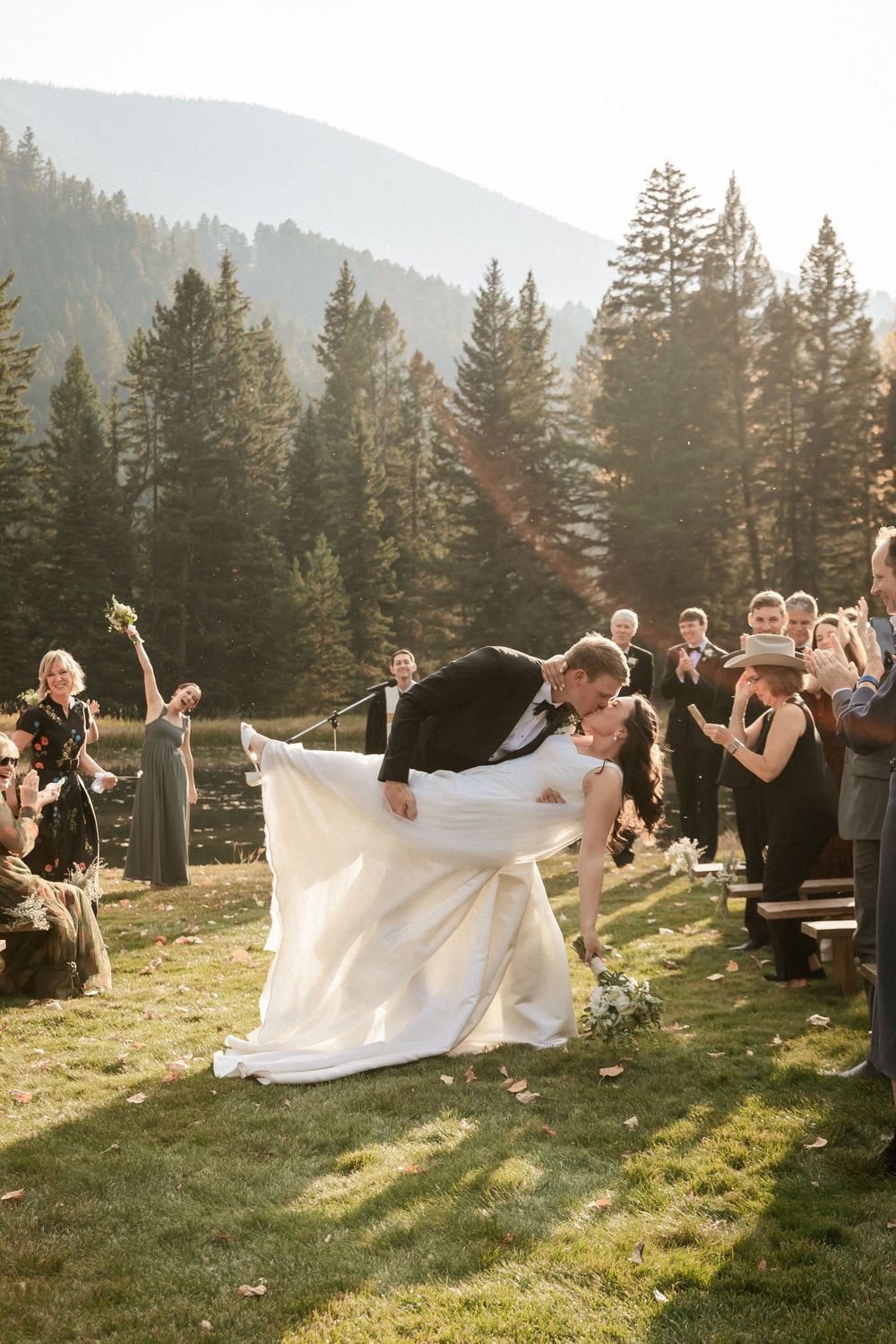 A couple shares a romantic dip during their mountain wedding ceremony as guests look on during golden hour.
