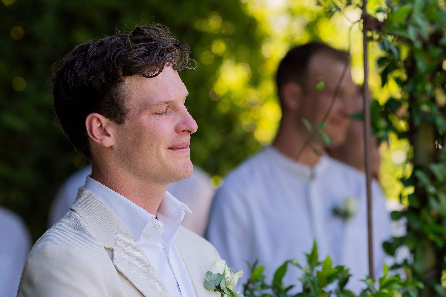 Groom in white suit jacket smiling during outdoor garden ceremony.