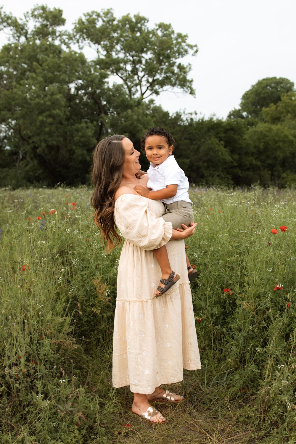 A tender moment shared between parent and child in a wildflower meadow with red poppies and lush green trees.