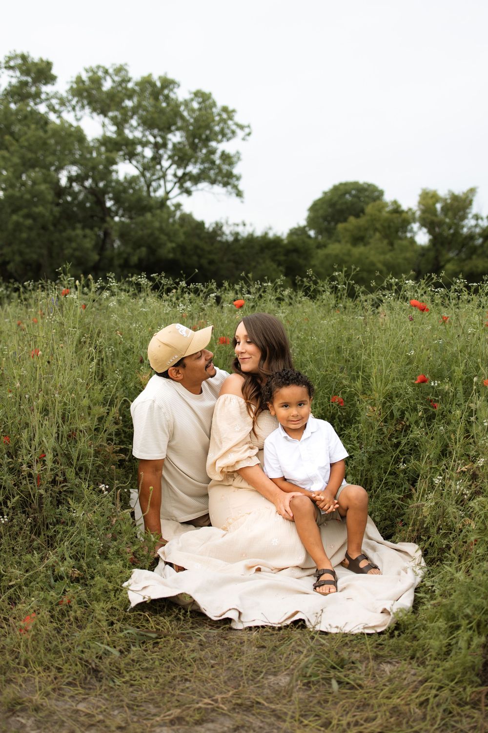 Family sitting together on blanket in meadow with wildflowers and red poppies on sunny summer day.