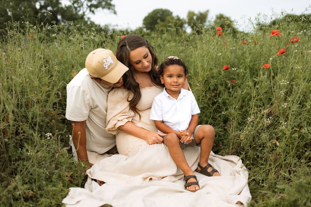 Three people sit together on a blanket in a wildflower meadow with red poppies blooming on a summer day.