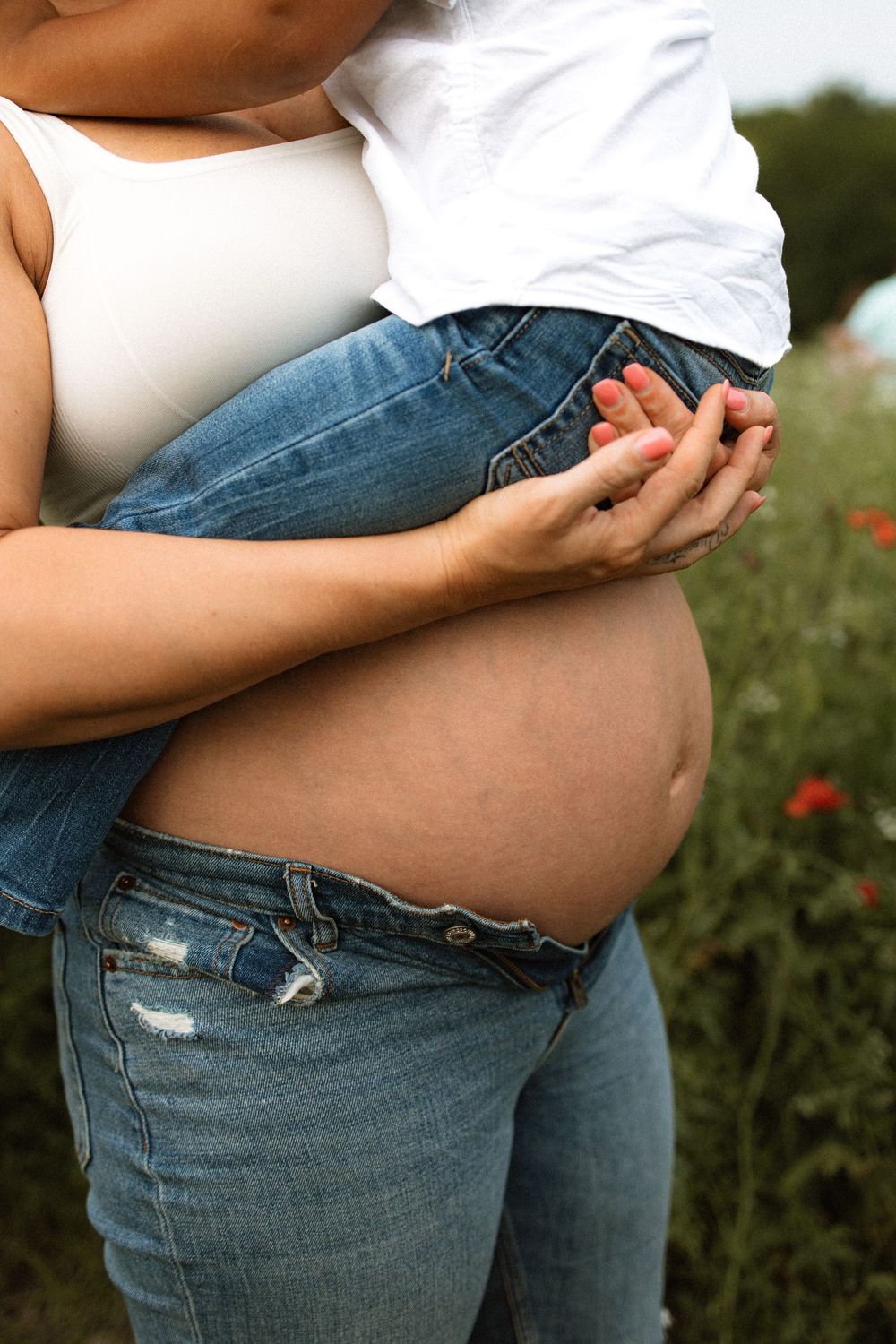 Close-up of expectant parents holding each other outdoors in a field with red flowers, wearing jeans and white top.