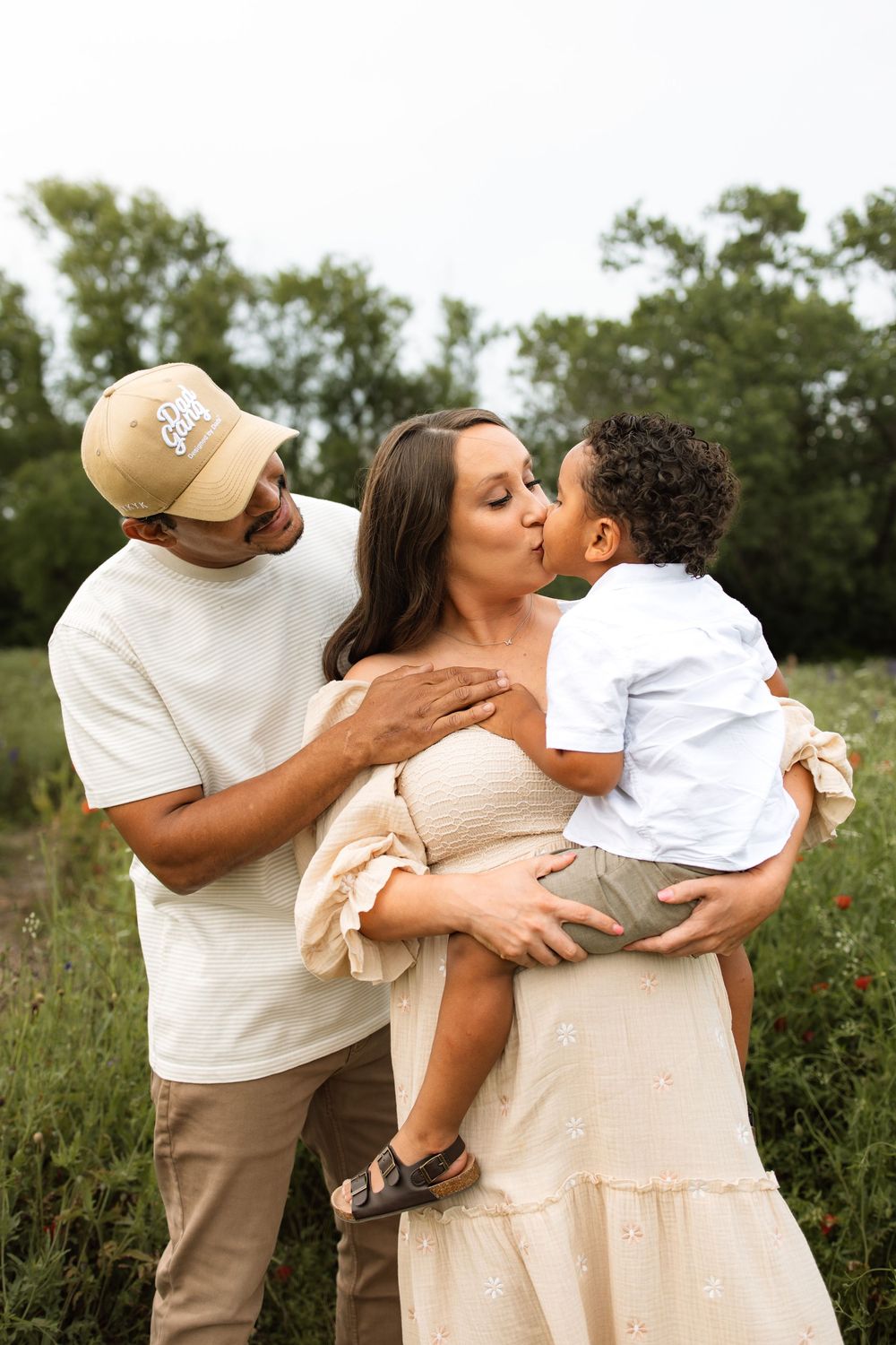 A family shares a tender moment together in a natural outdoor setting with tall grass and trees in the background.
