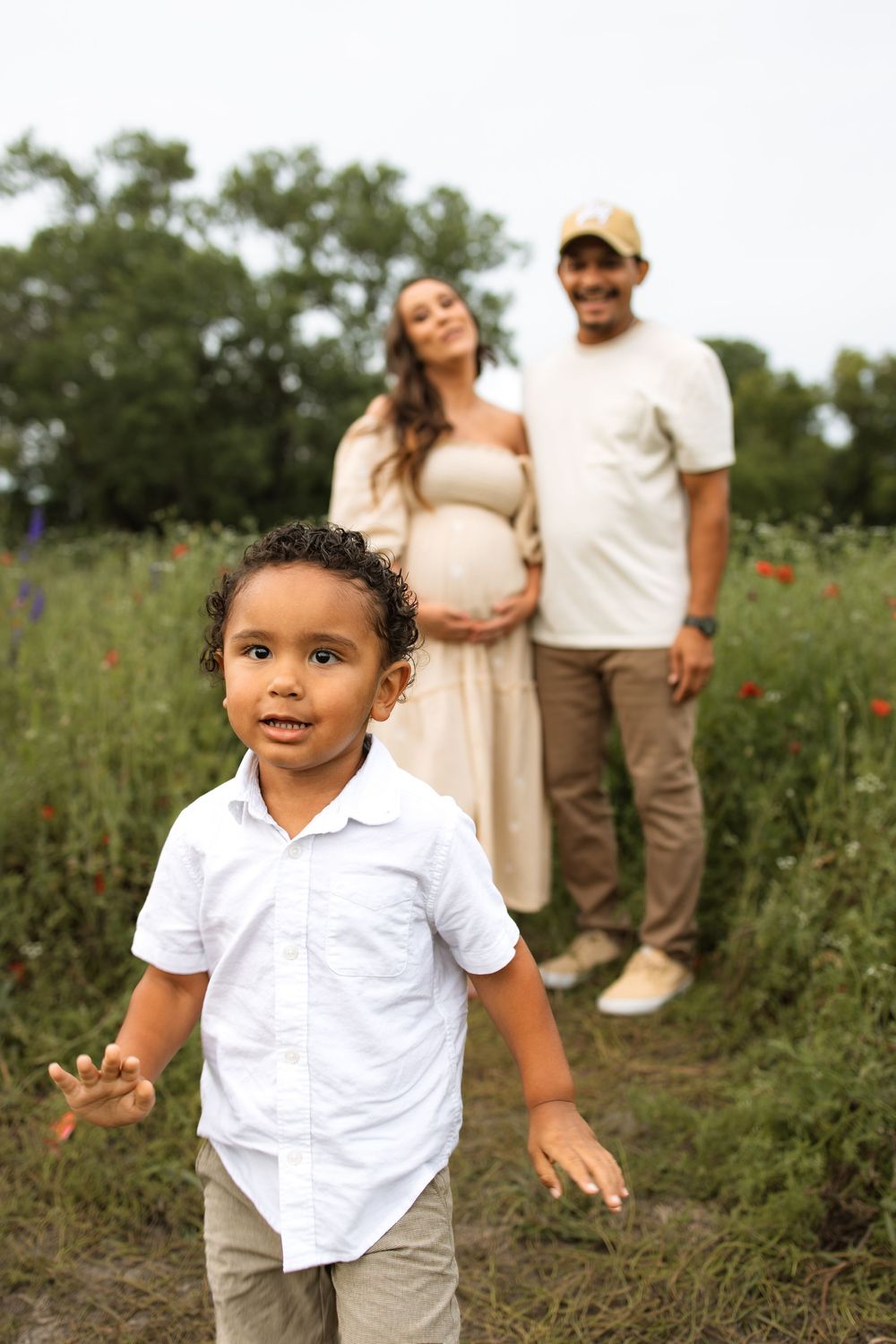 A young child in a white shirt joyfully plays in a wildflower field while a couple stands in the background.
