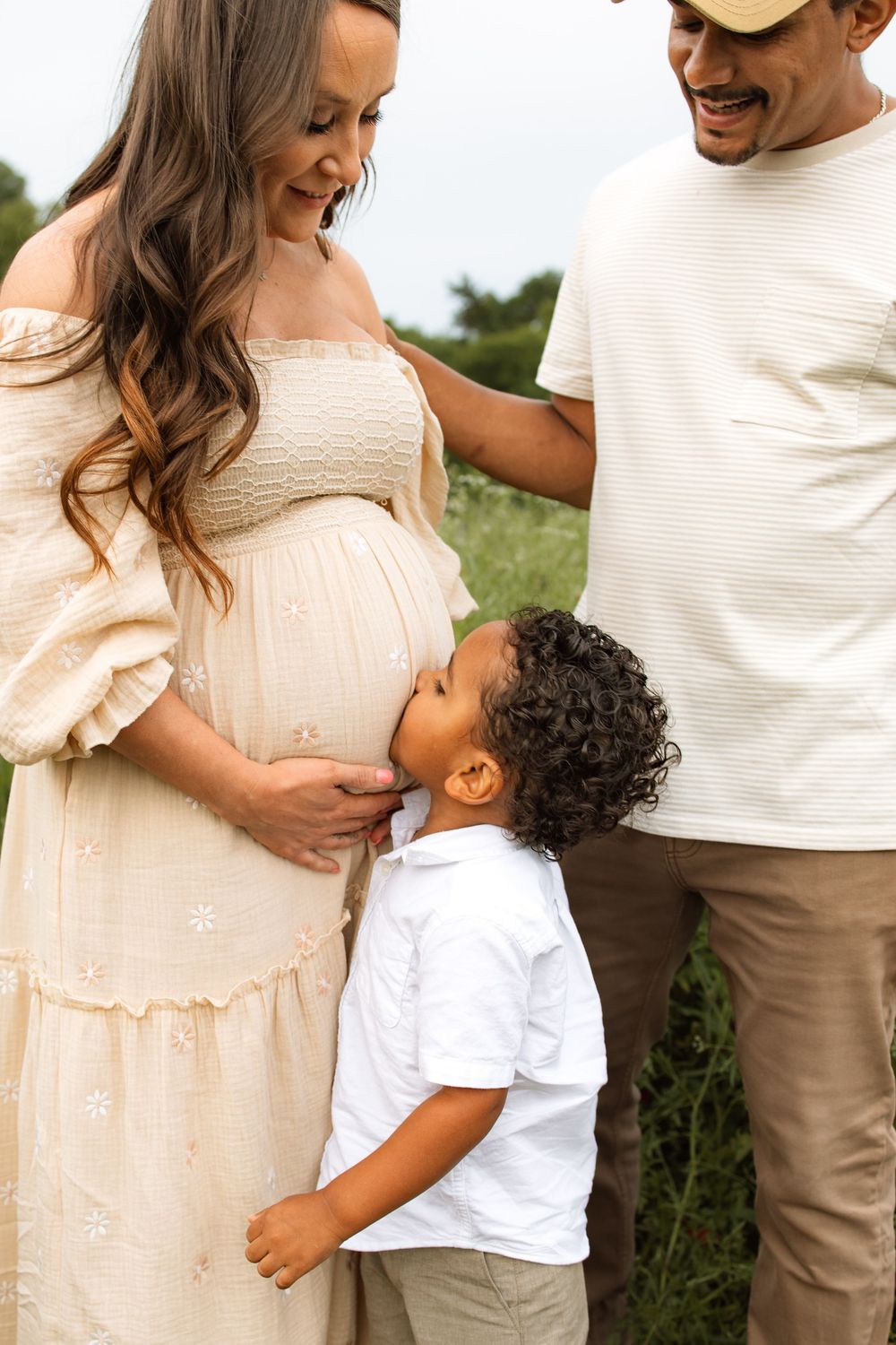 Family in outdoor setting with pregnant woman in cream dress, young child touching her belly, sharing tender moment in nature.