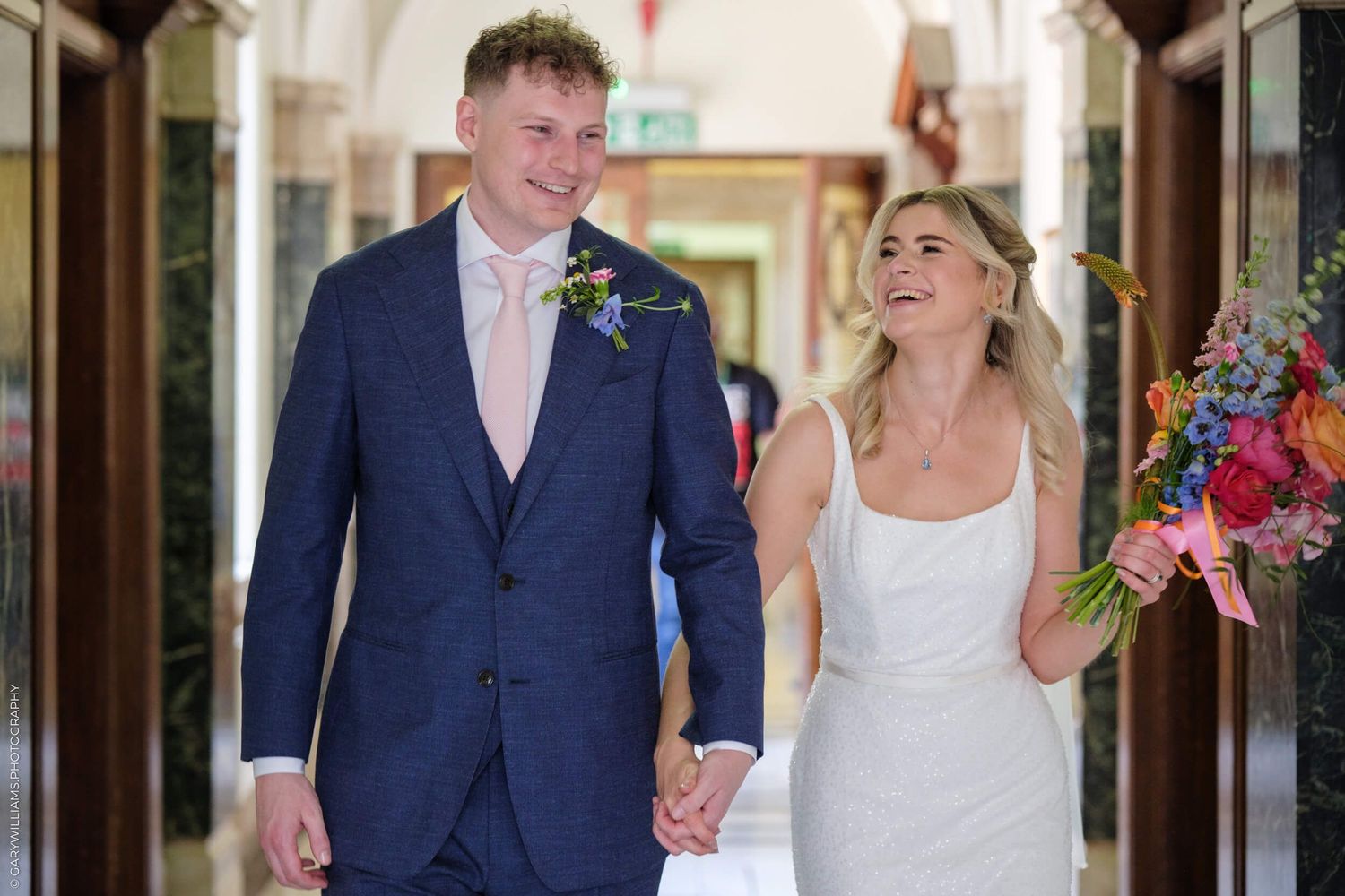 Newlywed couple walking down hallway holding hands and laughing joyfully in wedding attire and formal wear.