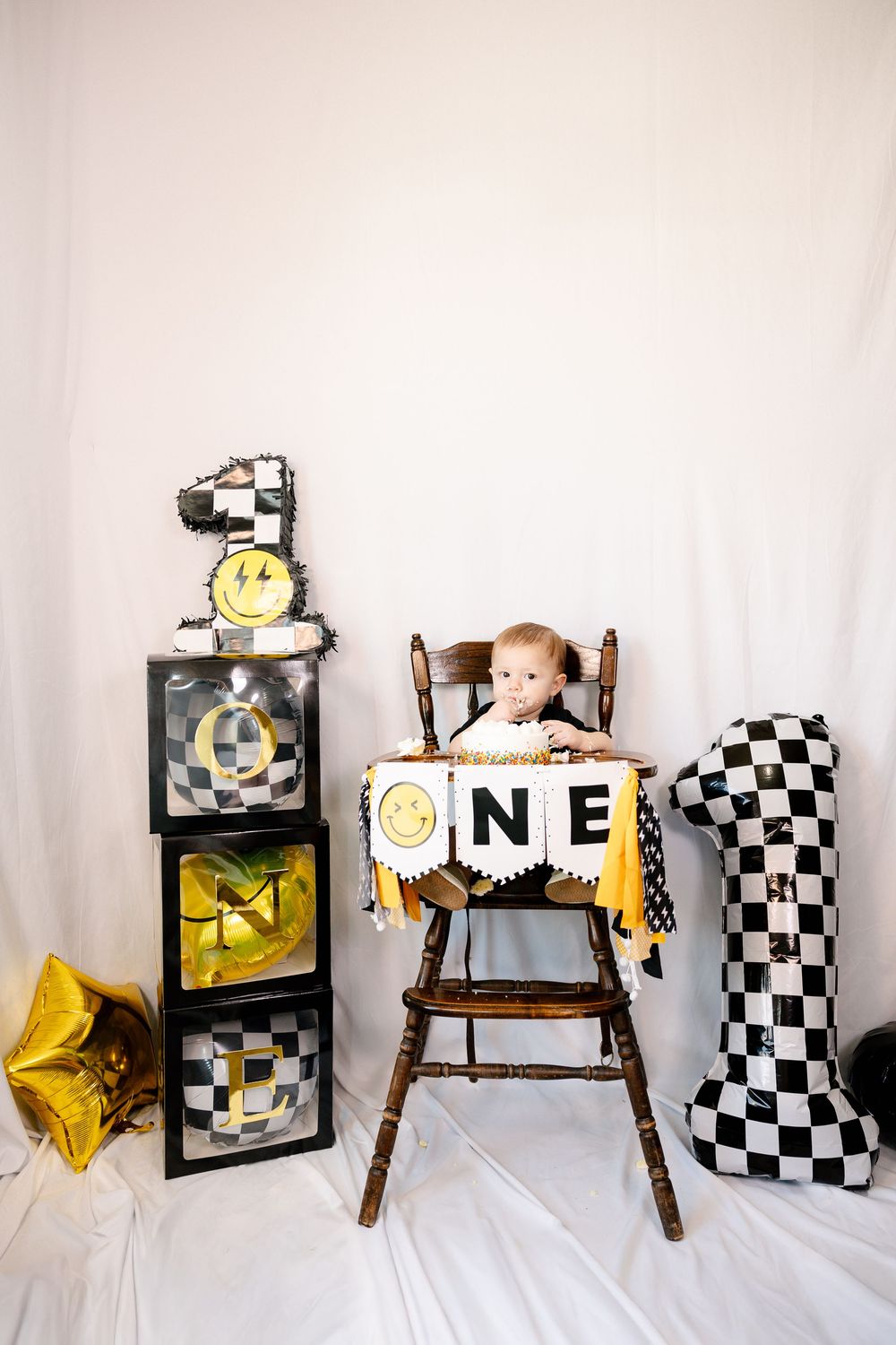 First birthday setup with wooden chair, checkered prints, and yellow star balloons against white background.