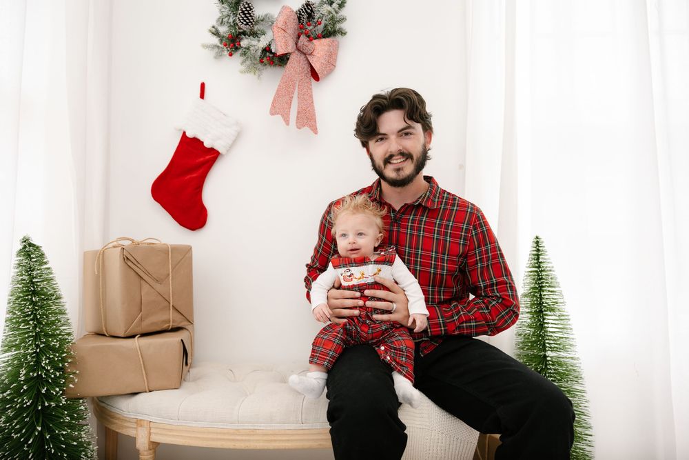Father holding baby in Christmas setting with stockings, gifts, and holiday decor in a white studio.