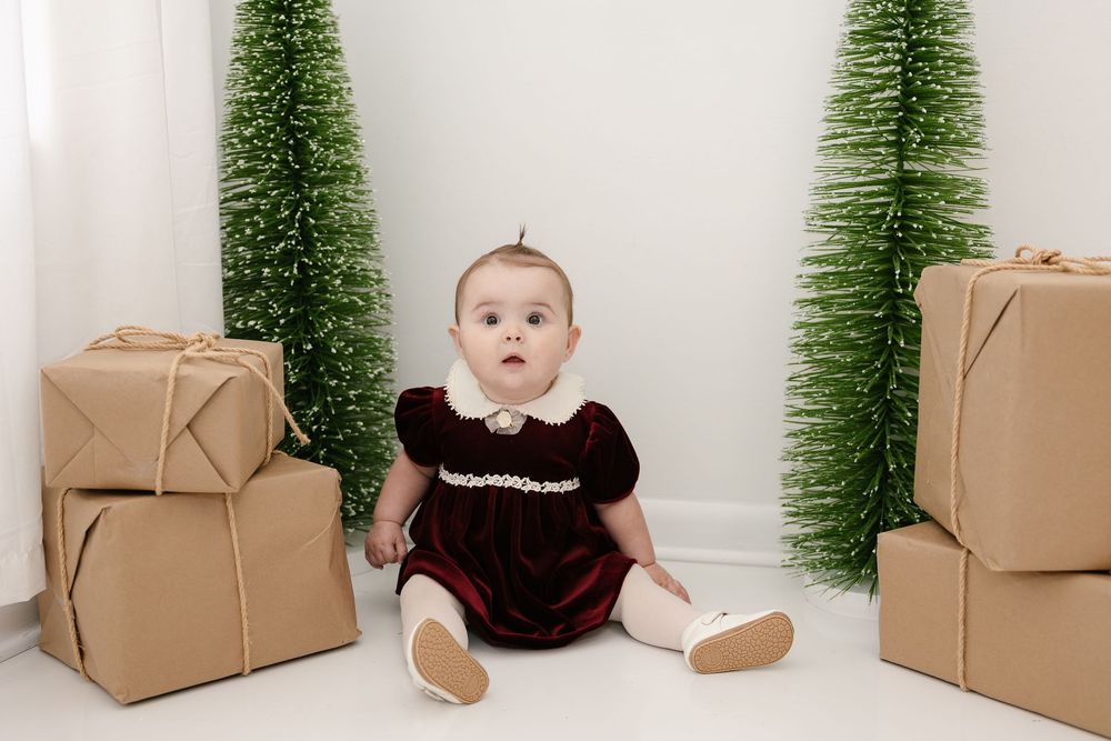 Baby in red velvet dress sits between wrapped gifts and green bottle brush Christmas trees.