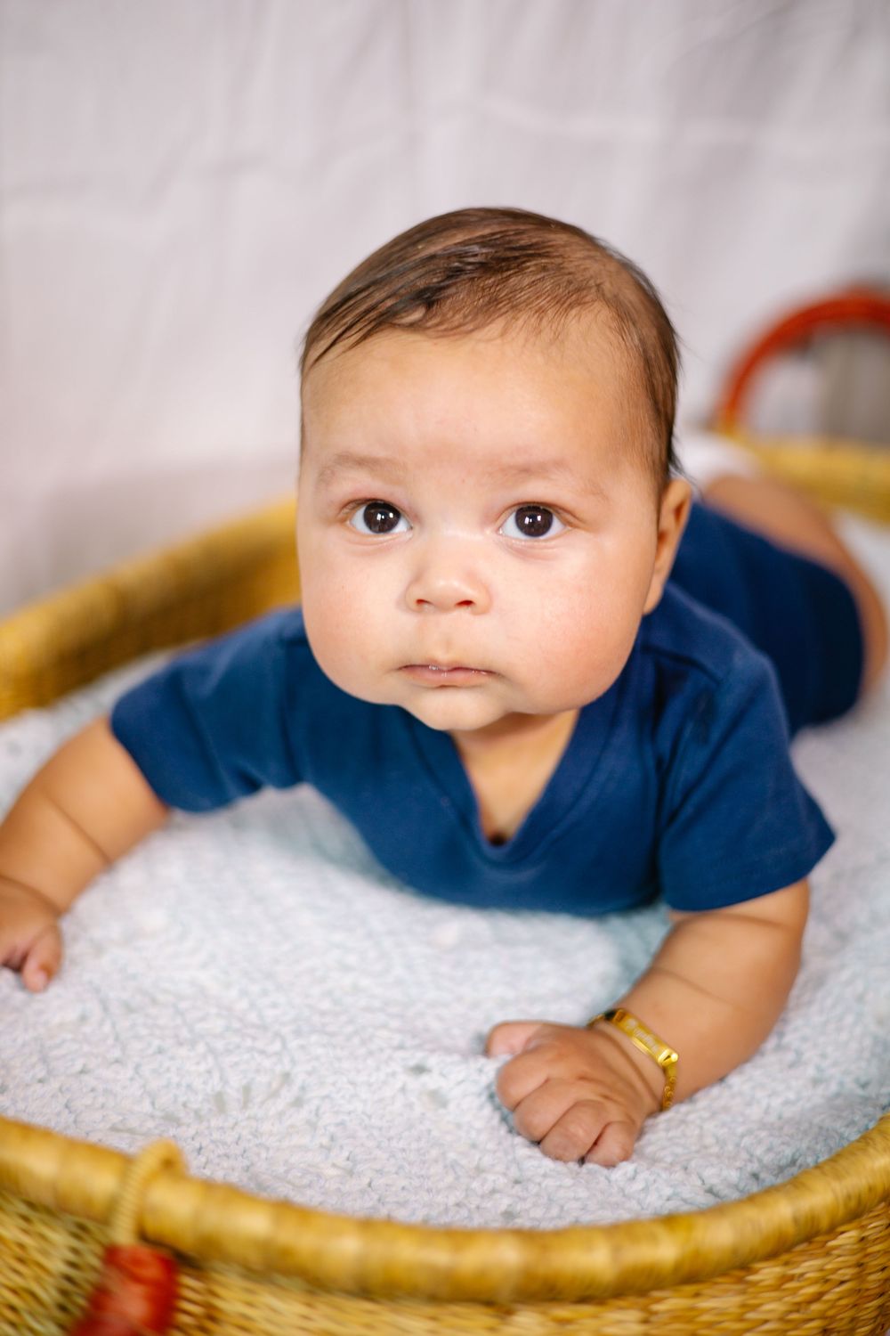 Adorable baby in blue onesie lying in a wicker basket on a soft white blanket, looking up with curious brown eyes.