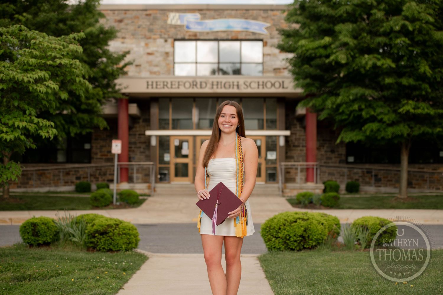 Graduation photo taken outside traditional brick high school building entrance with landscaped shrubs and greenery.
