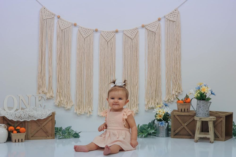 Small child sits on floor with macrame backdrop, floral decor and wooden crates creating a boho photography setup.