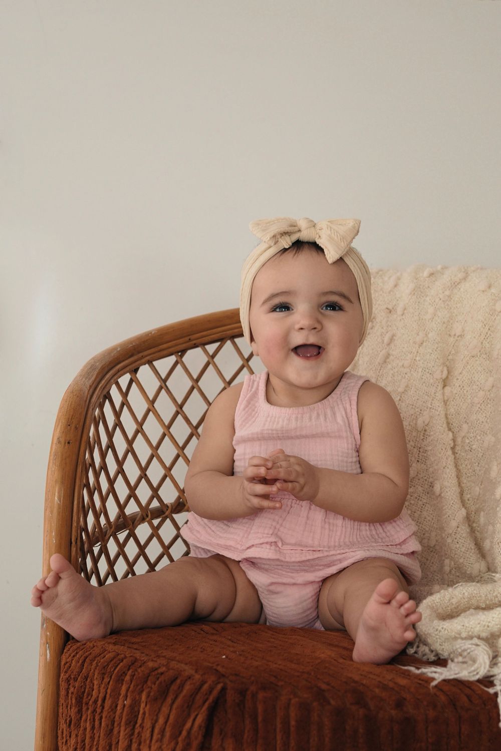 A happy baby wearing a pink romper and beige headband sits in a woven rattan chair with a cream blanket.