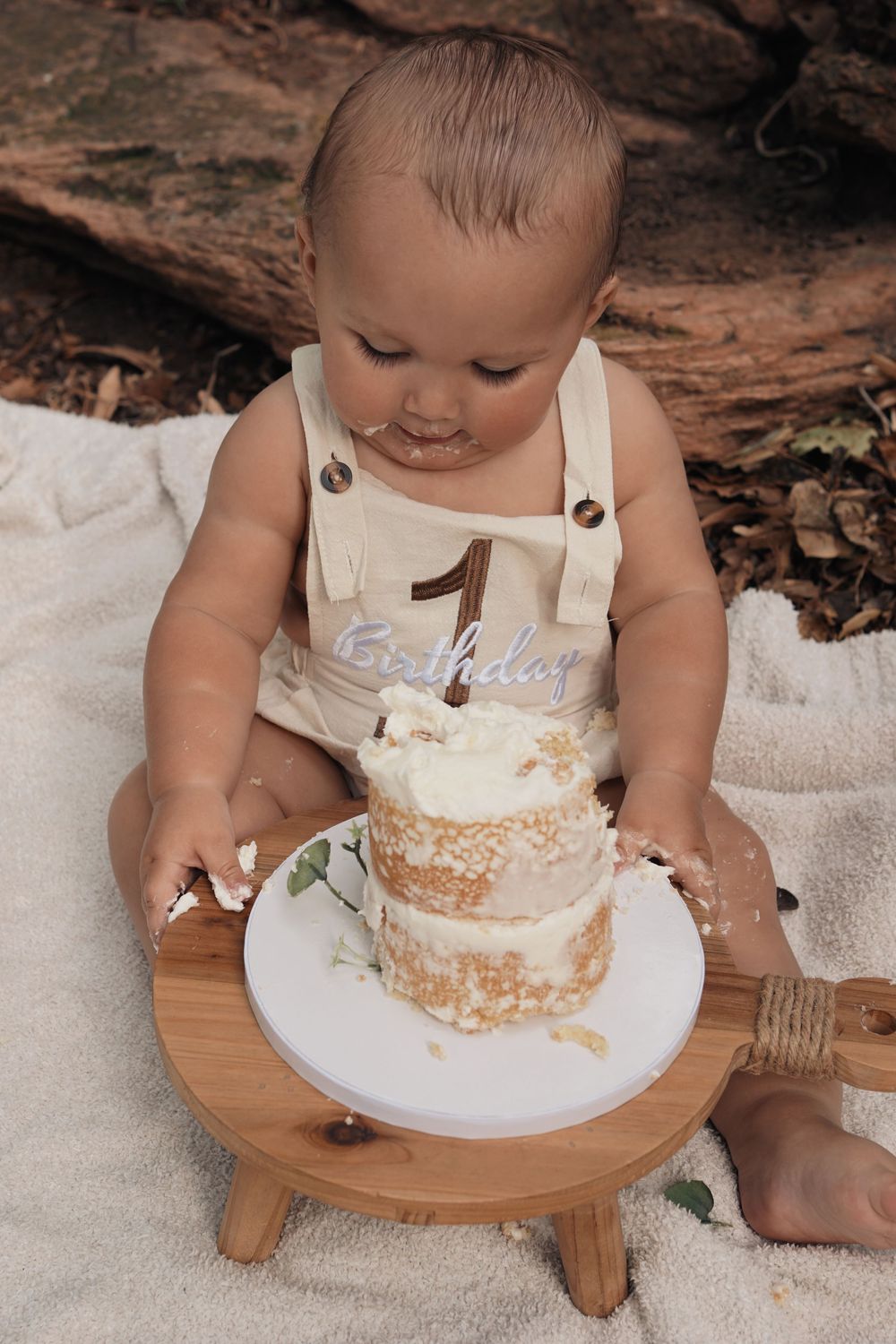 Baby in white overalls sits at wooden stool enjoying their first birthday cake smash photoshoot on white blanket.
