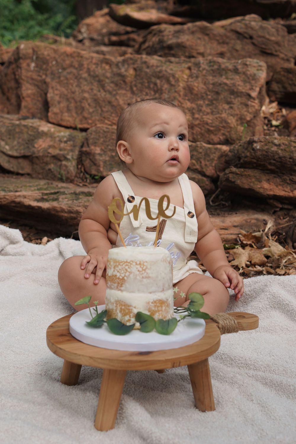 A one year old sits on a white blanket with a small birthday cake on a wooden stool against a rock wall background.