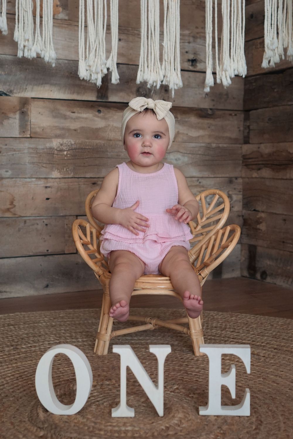 A small child in a pink romper sits on a wooden chair with macrame decor and white ONE letters for first birthday photo.