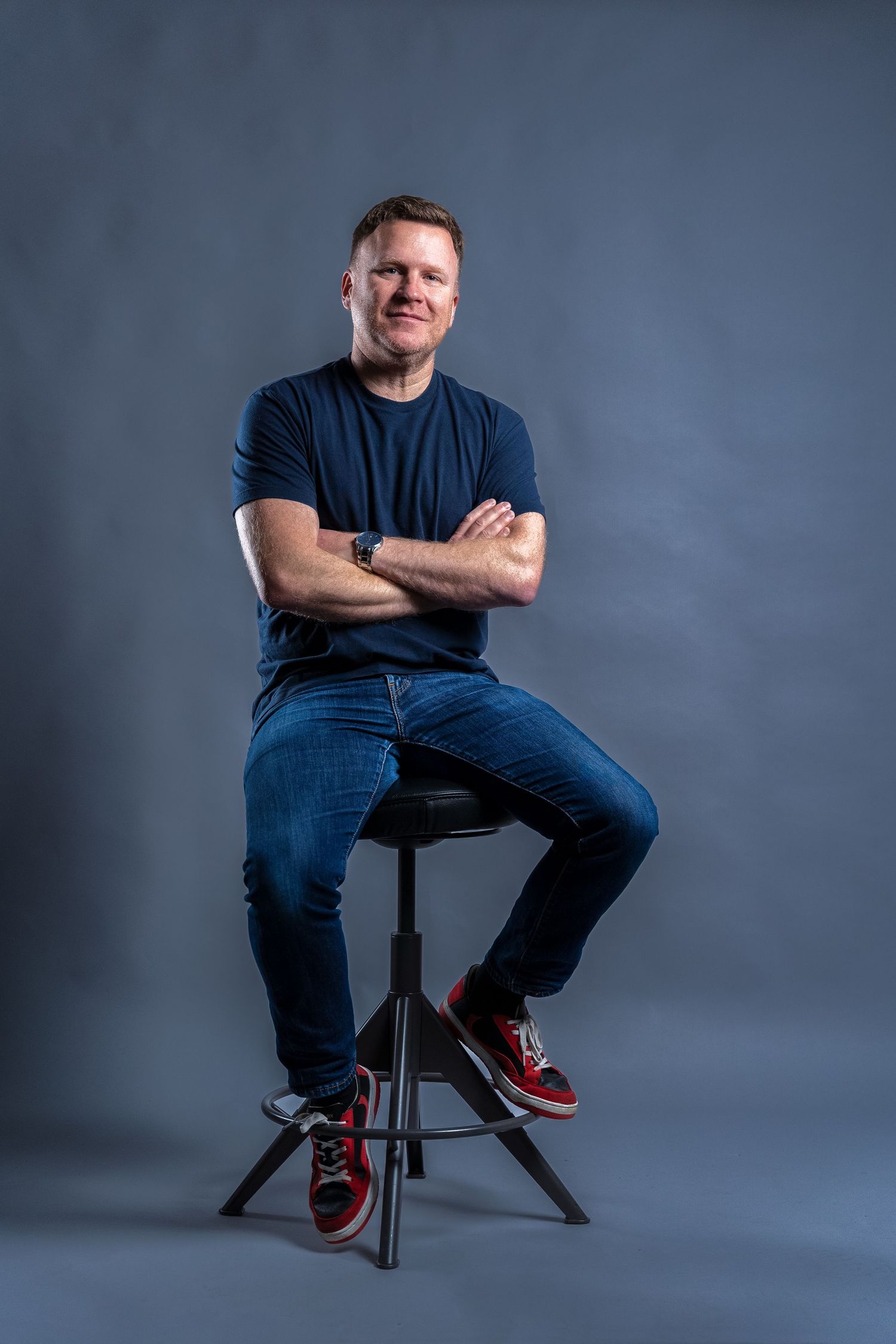 Entrepreneur wearing navy t-shirt and jeans sits casually on black stool against grey on a personal branding studio photoshoot