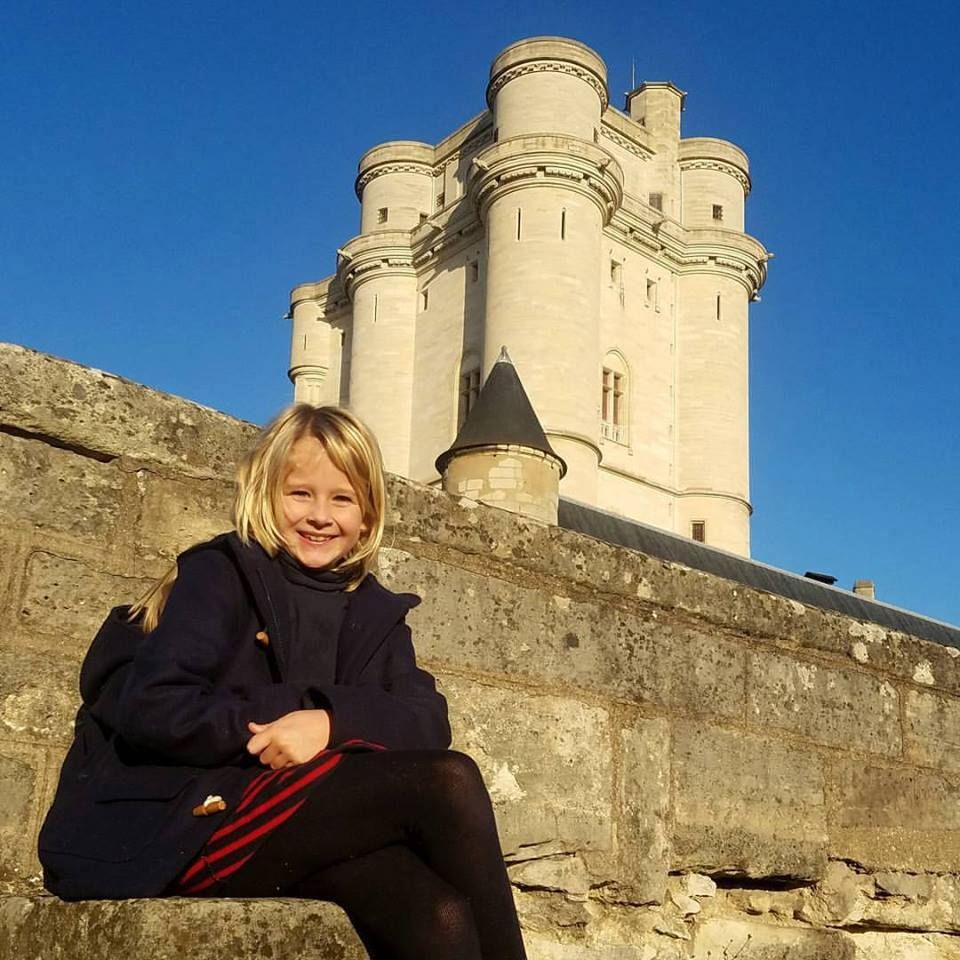 Medieval castle towers rise against bright blue sky as visitor poses on ancient stone walls.