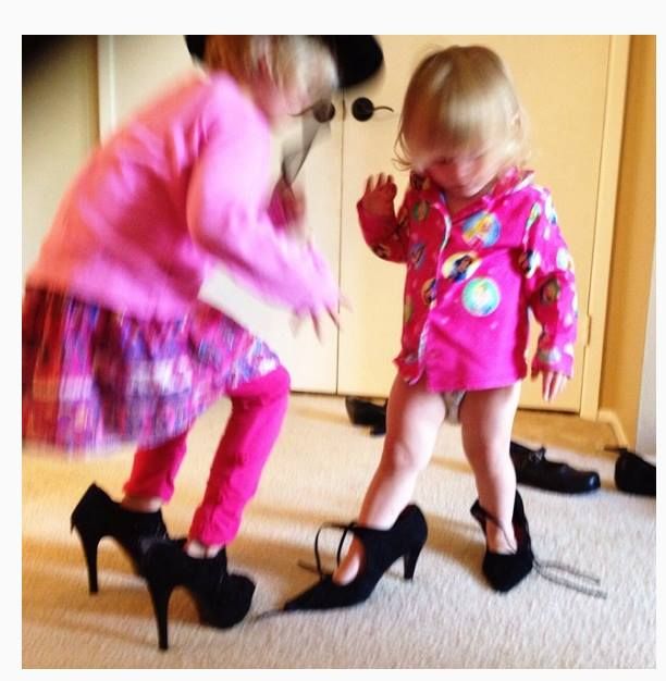 A small child in pink clothing attempts to walk in oversized high heel shoes on a beige carpet.