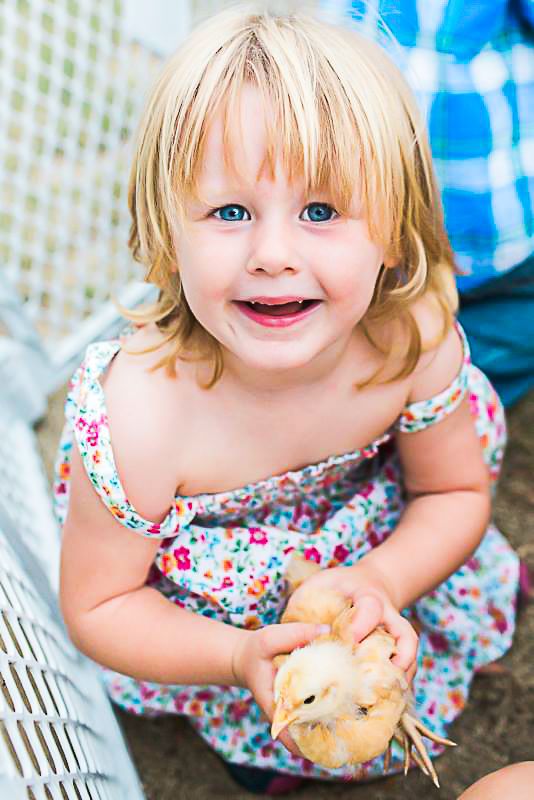 A young child in a floral dress holds a baby chick while smiling brightly.
