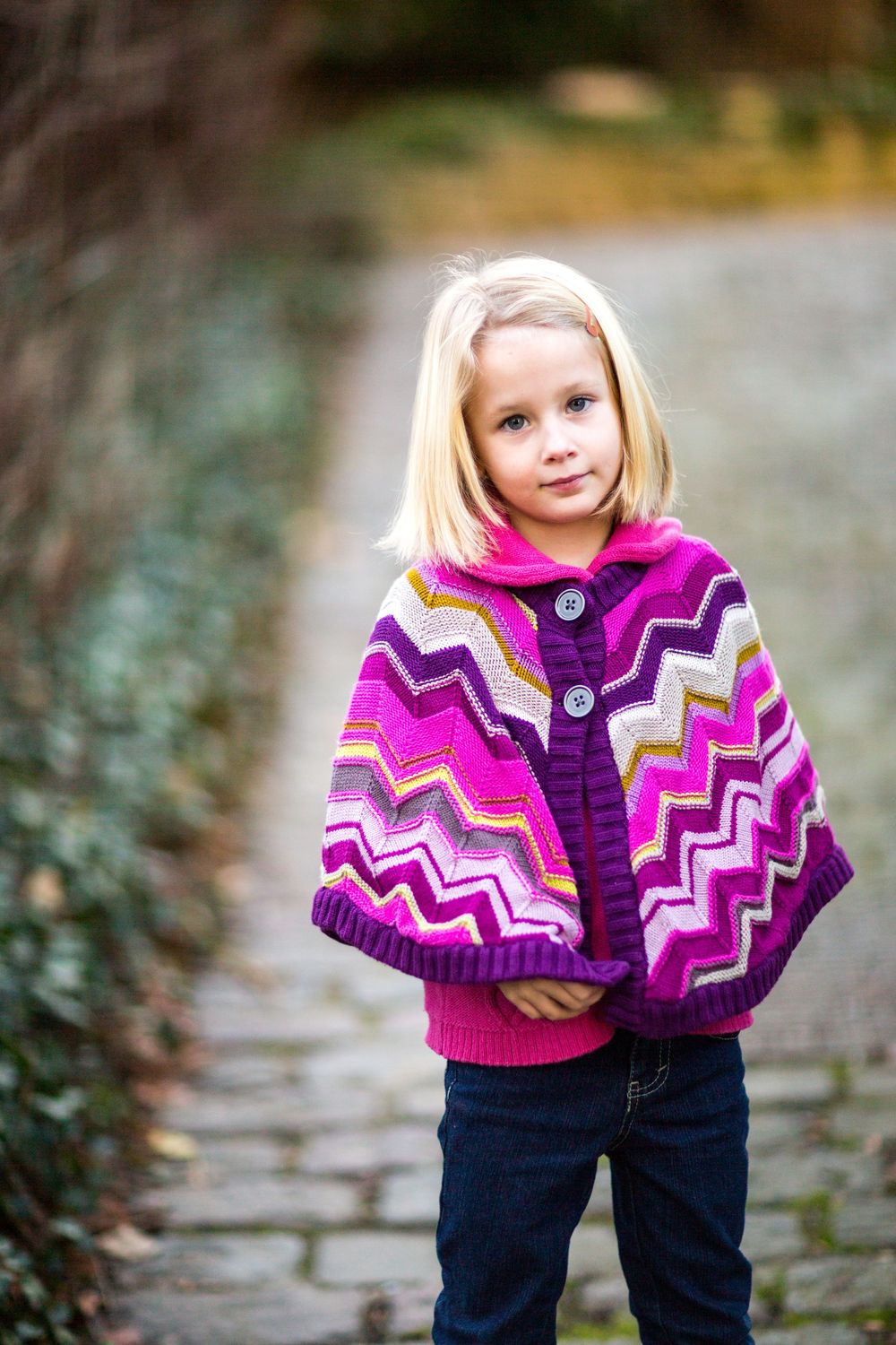 A child wearing a vibrant knitted poncho with purple and pink chevron patterns stands on a stone path.