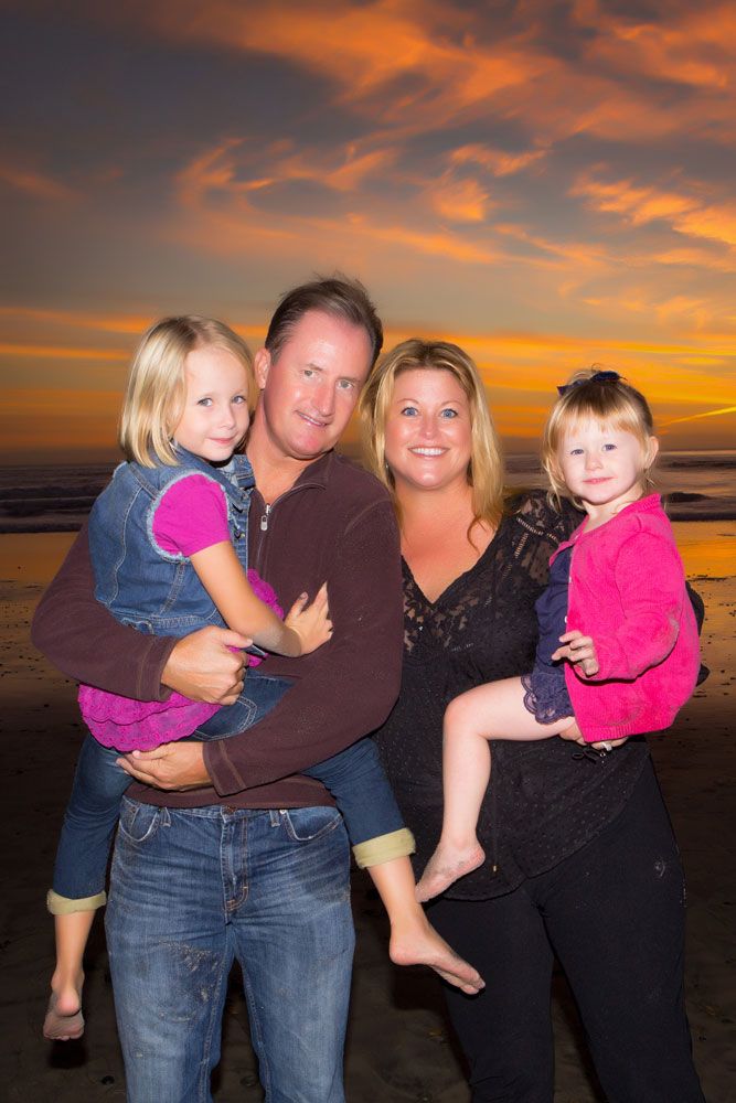 A family poses together on a beach at sunset with dramatic orange and pink clouds in the background.