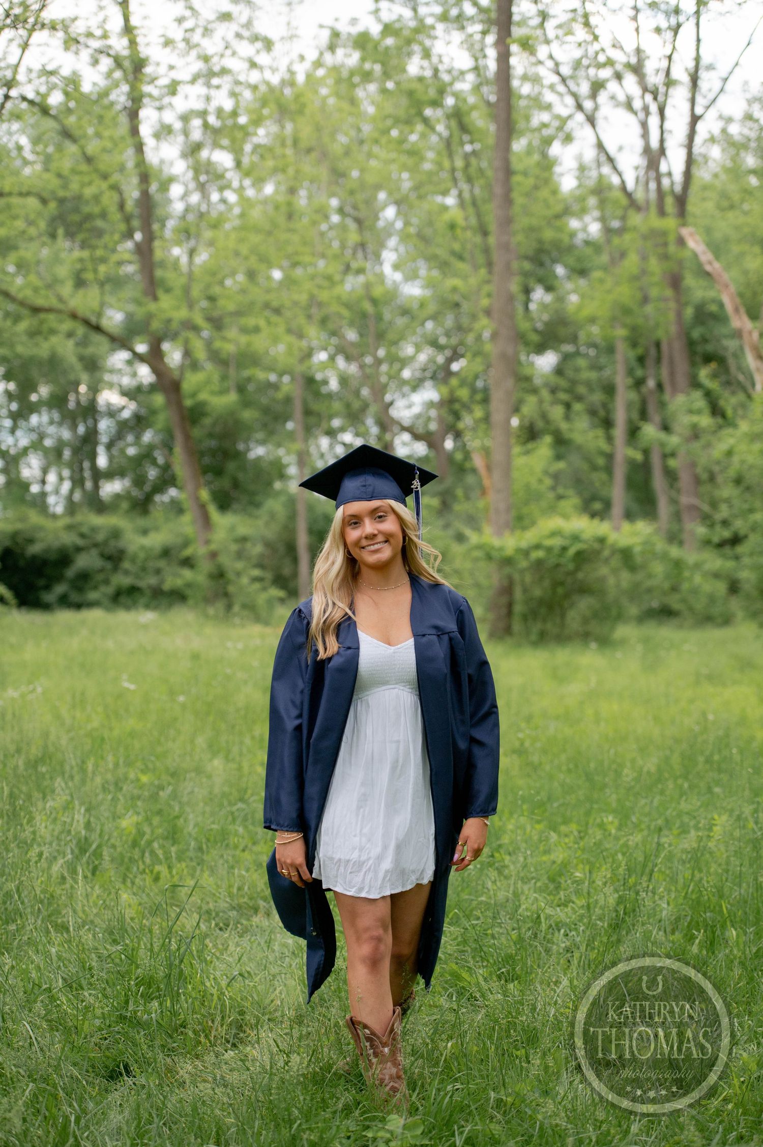 A graduate posing in an outdoor grassy field wearing graduation regalia and white dress for photos in nature.
