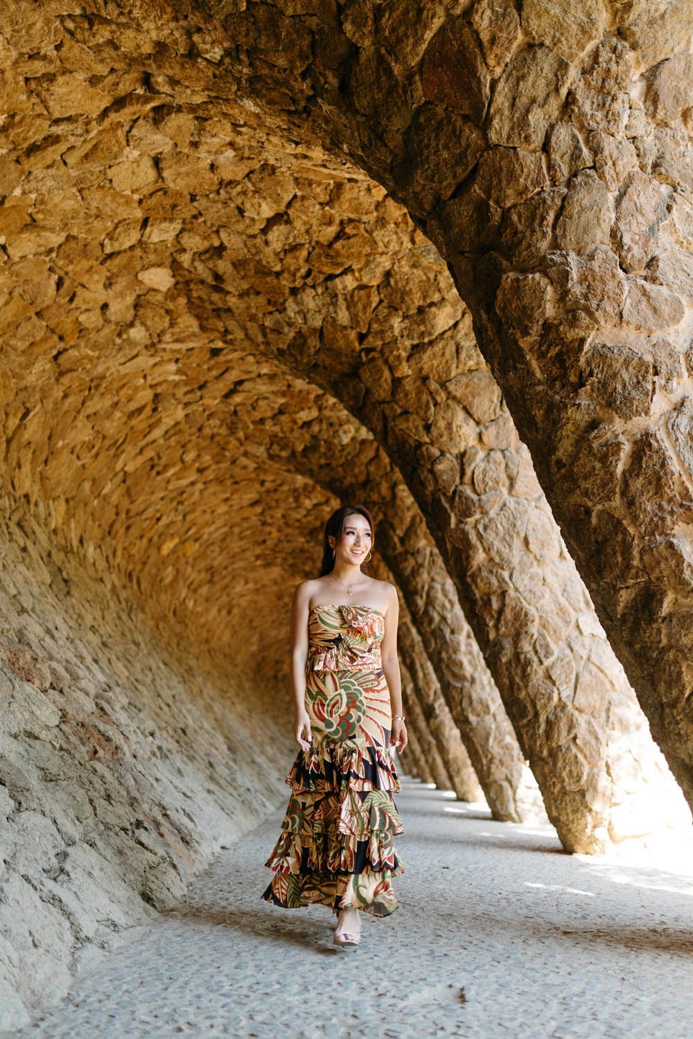 Woman in floral dress poses under stone archways in Park Guell, Barcelona.