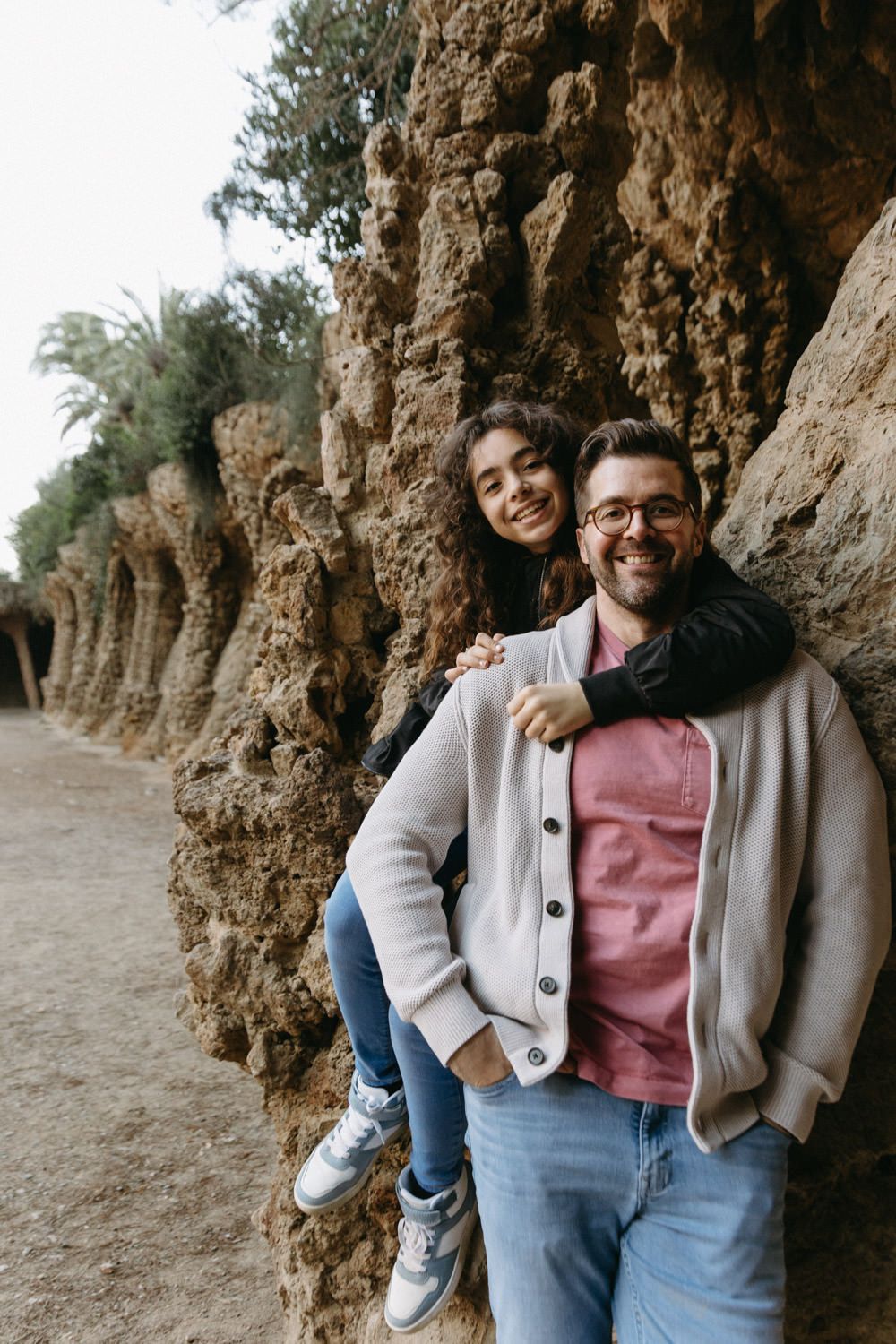 Couple poses casually against rustic stone wall in park setting.