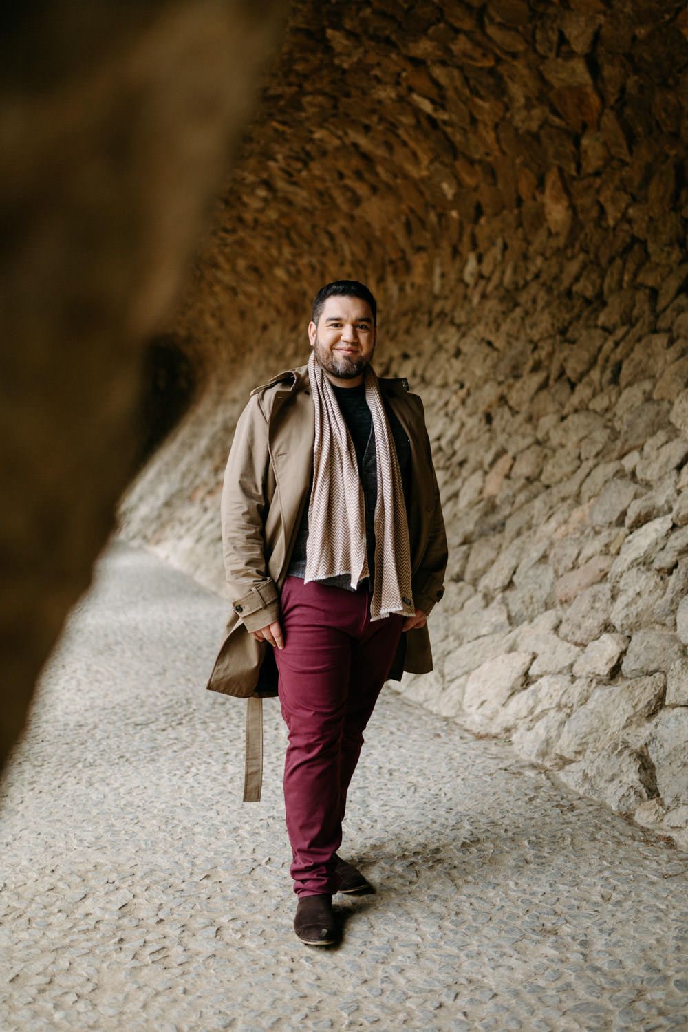 Person walking through curved stone tunnel wearing brown coat and burgundy pants.
