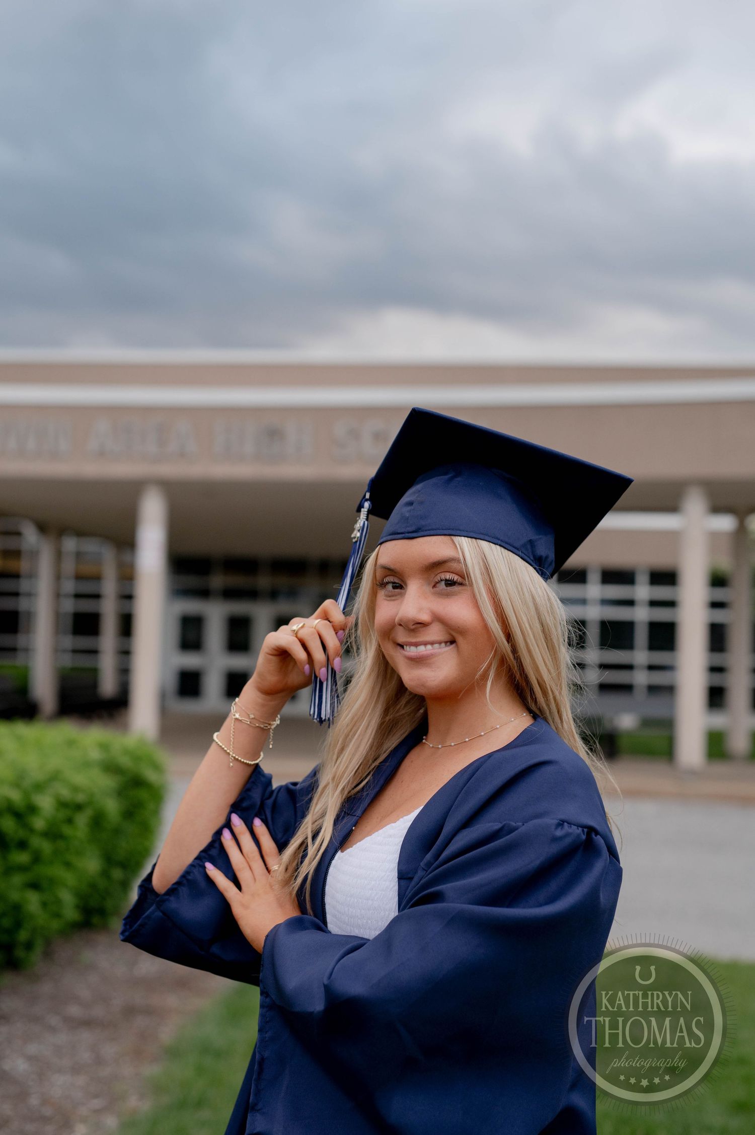 A graduate in navy cap and gown poses outdoors in front of a school building on a cloudy day.