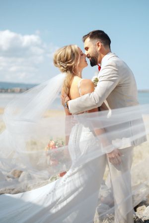 Romantic beach wedding portrait with couple's veil flowing in ocean breeze.