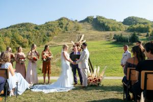 Outdoor mountain wedding ceremony with bridal party standing on grassy hillside during sunset.