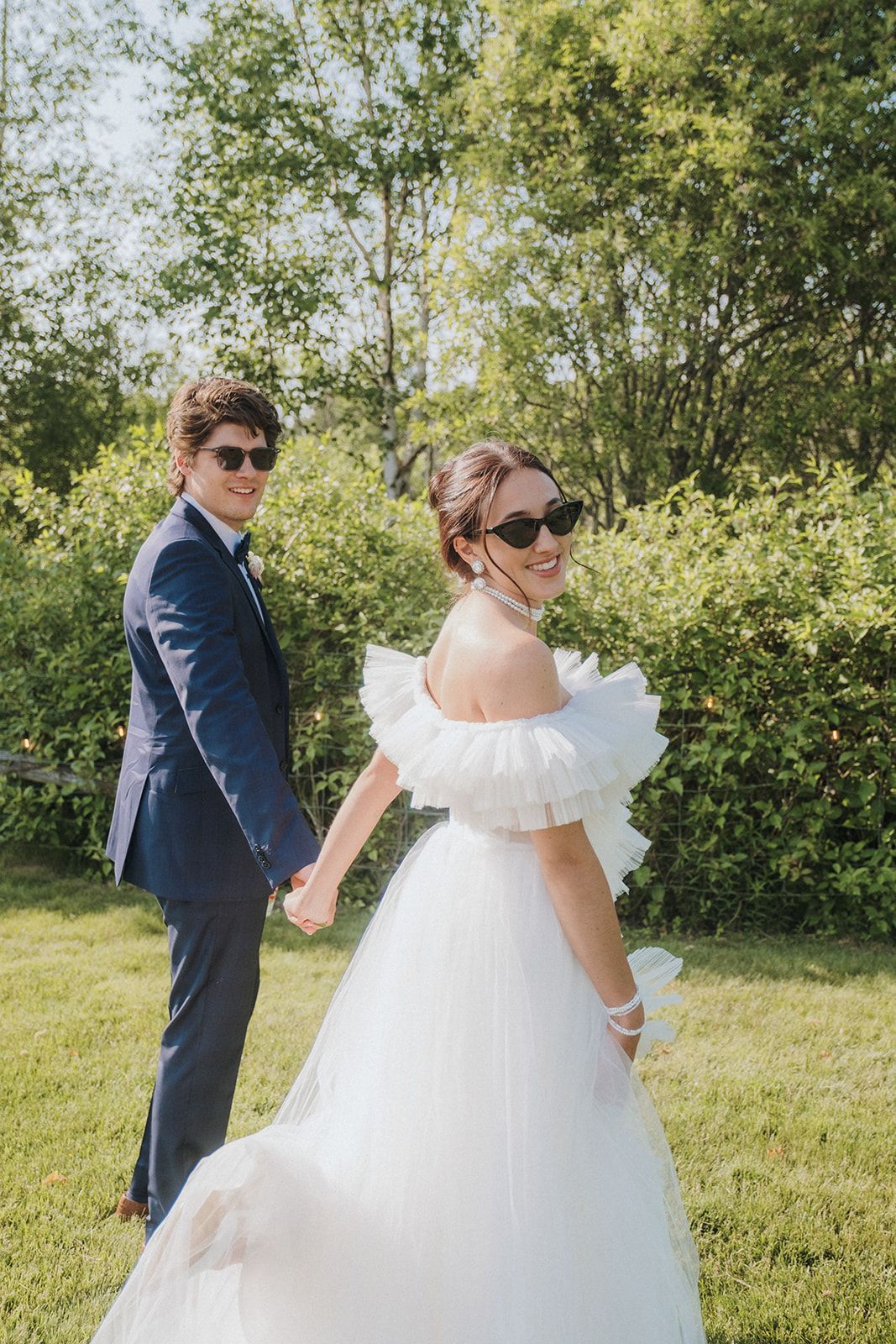 Stylish couple wearing sunglasses pose in garden setting with bride in off-shoulder white dress.