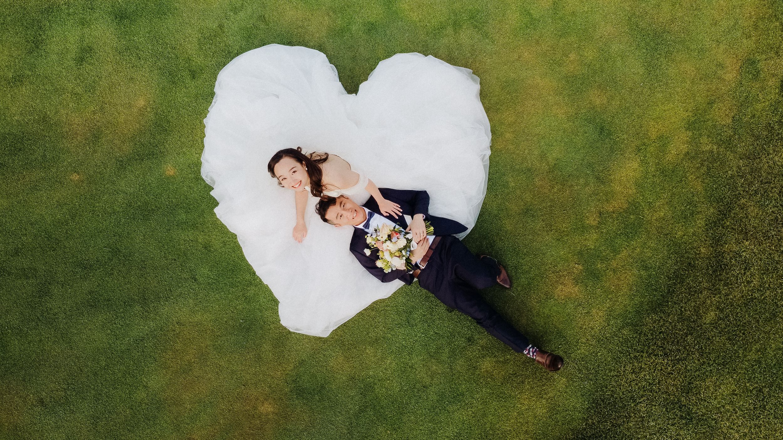 Aerial view of a couple lying on grass creating heart shape with wedding dress.