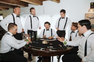 Groomsmen in white shirts and suspenders sharing drinks and laughs at a rustic wooden bar.