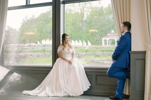 Wedding couple poses elegantly by large windows in formal attire.