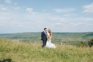 Couple stands on hilltop meadow with sweeping valley views in background.