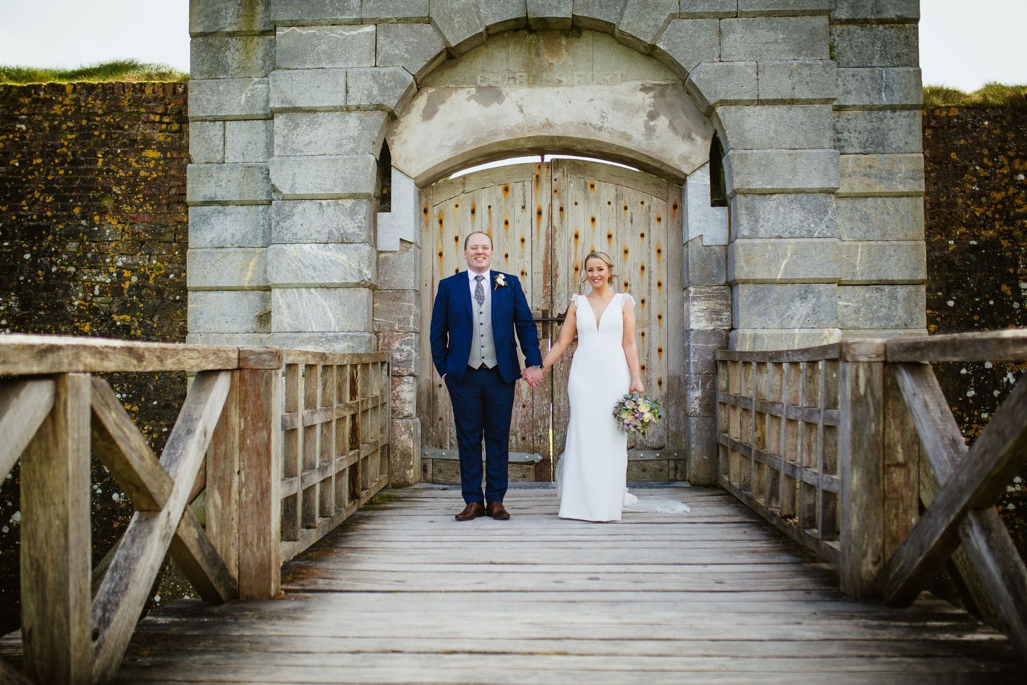 A couple stands on a wooden bridge under a stone archway at a historic fort entrance.