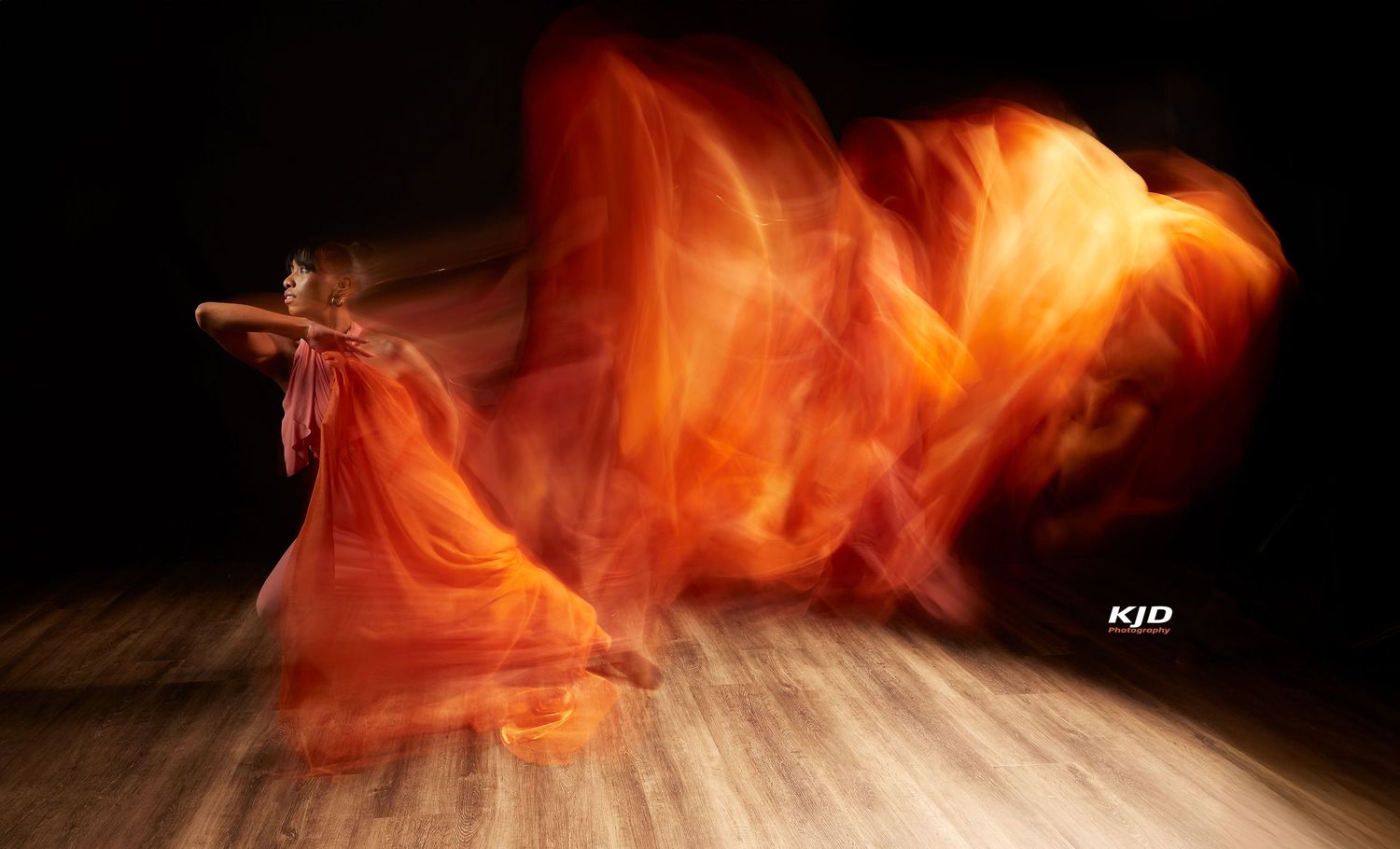 Long exposure photograph captures flowing orange fabric movement in dance creating flame-like motion trails on wooden stage.