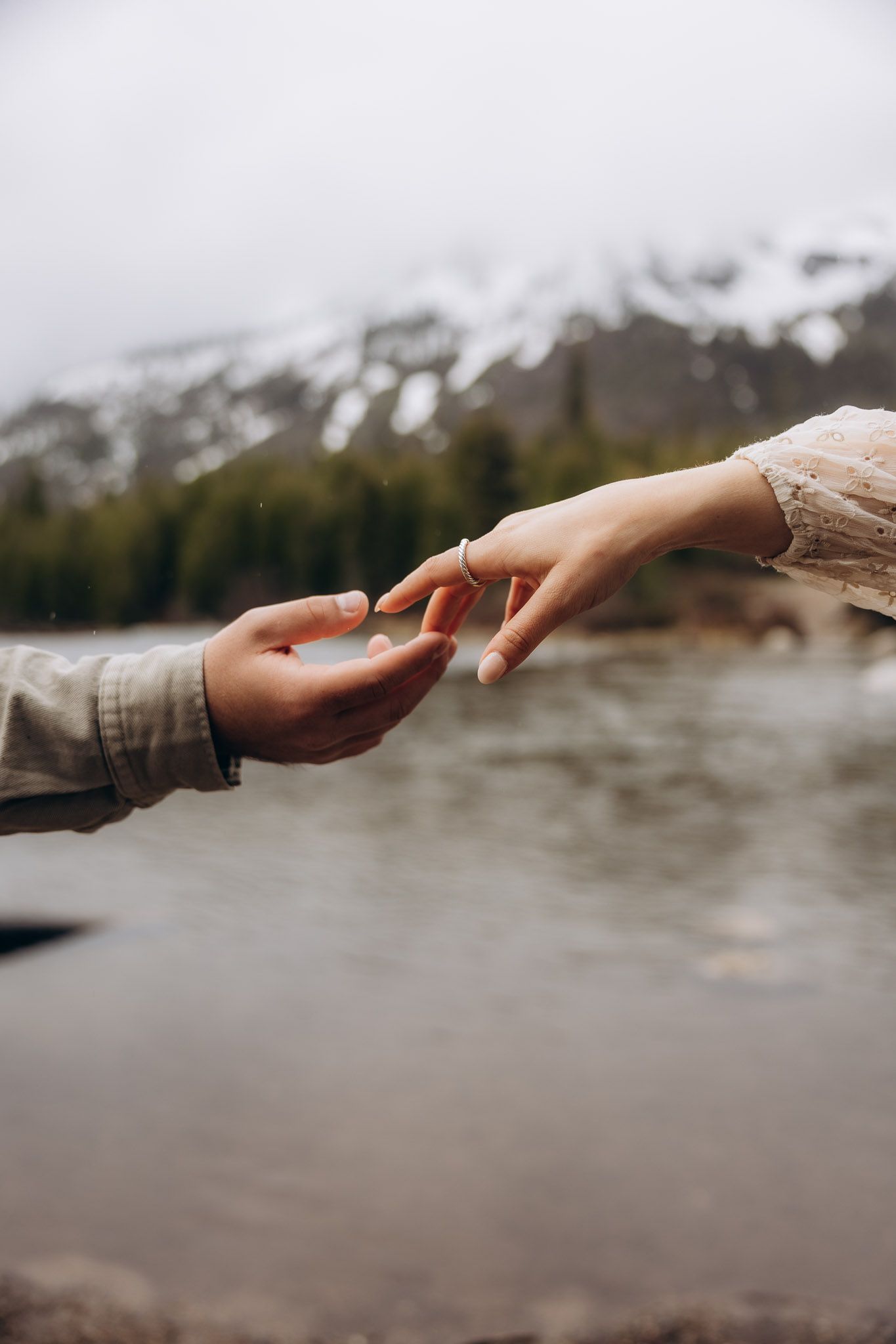 Two hands reaching towards each other against a backdrop of snowy mountains and a lake in moody lighting.