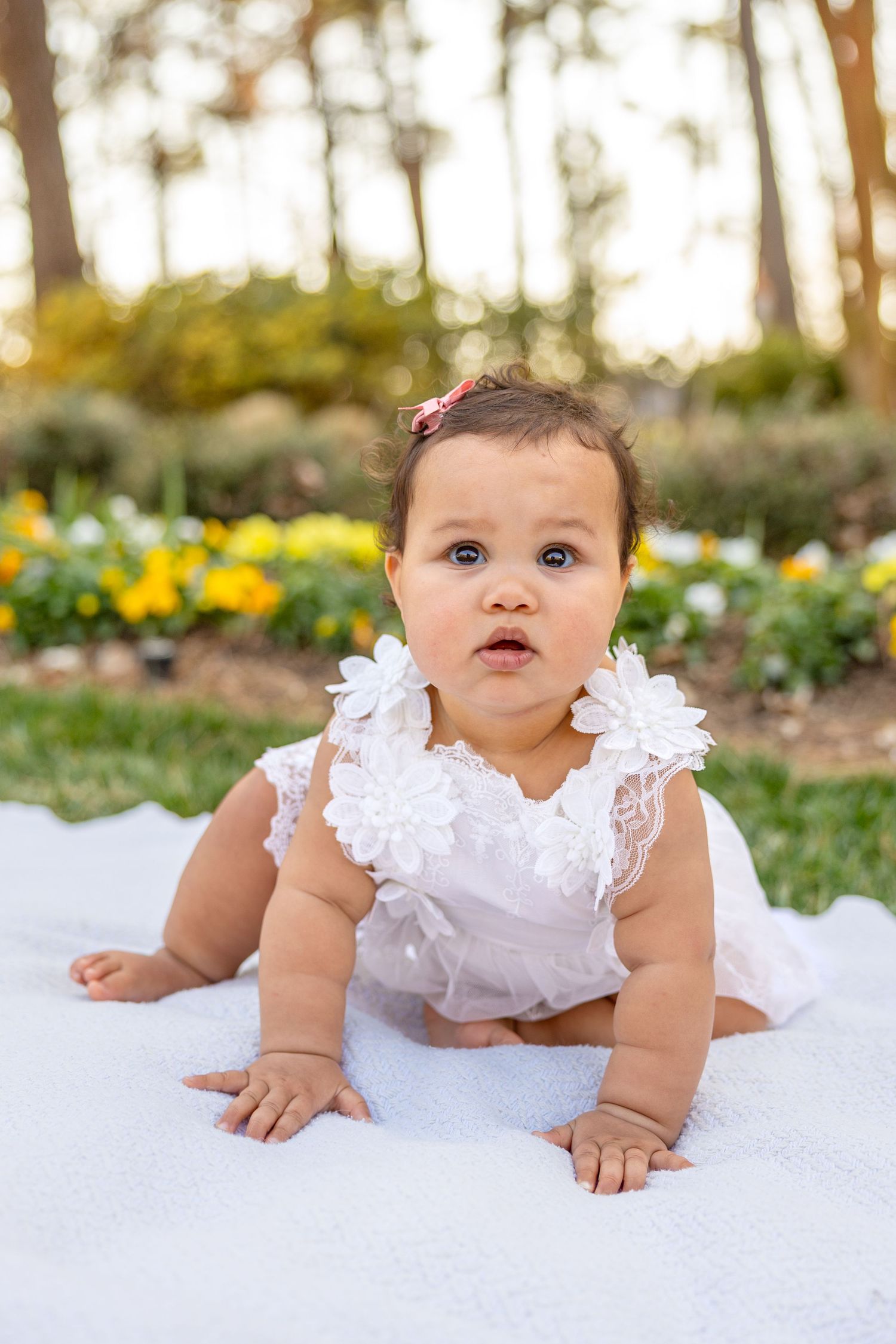 Baby wearing white lace dress lies on white blanket outdoors with yellow flowers and trees in background.