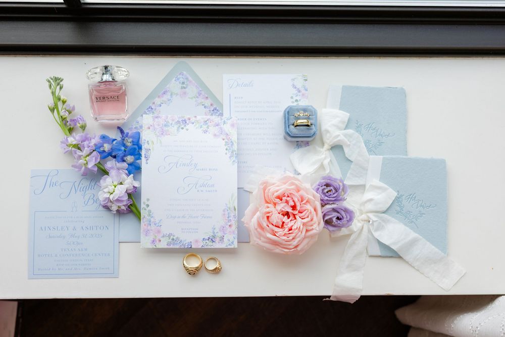 Wedding stationery flat lay with blue and pink flowers, ribbons, and perfume bottles on a white surface.
