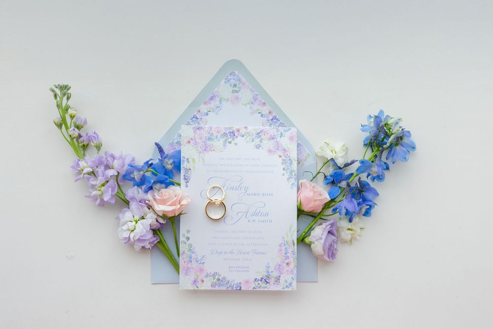 Wedding invitation card decorated with delicate blue and pink flowers and gold ring on white background.