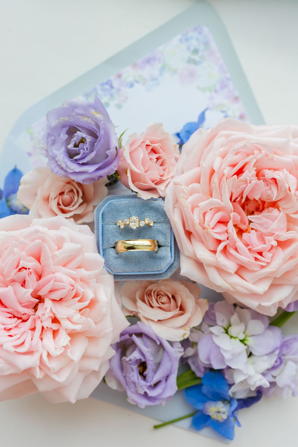 A delicate engagement ring in a blue velvet box surrounded by pink and purple roses and floral arrangements.