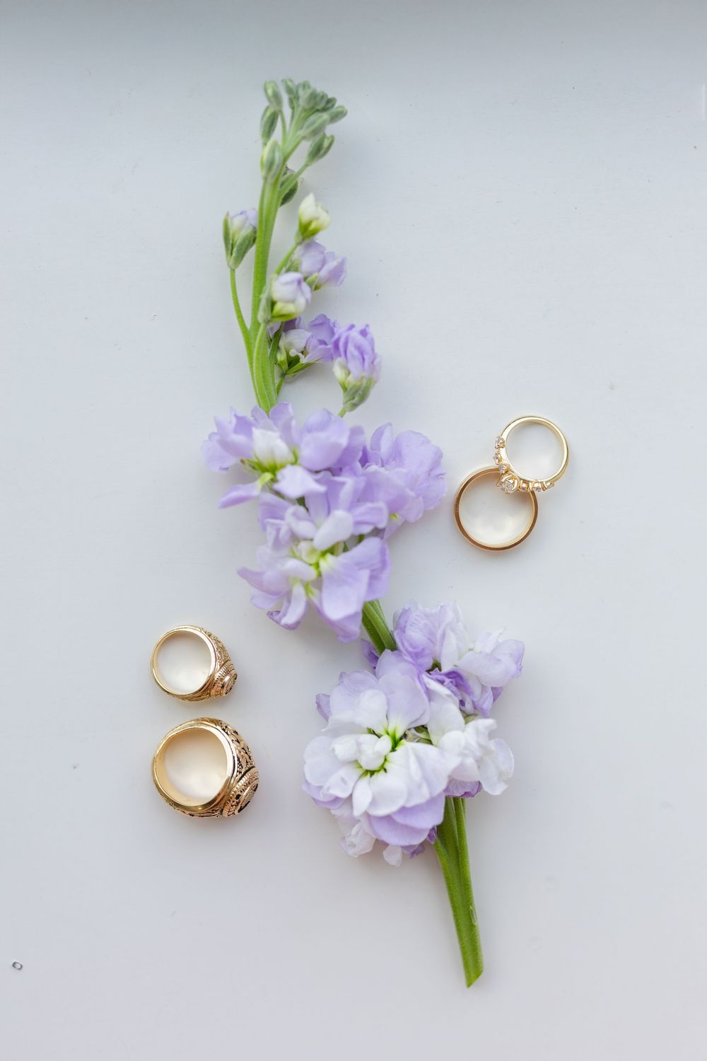 Delicate purple flowers arranged with gold wedding rings on a light gray surface for a romantic wedding detail shot.