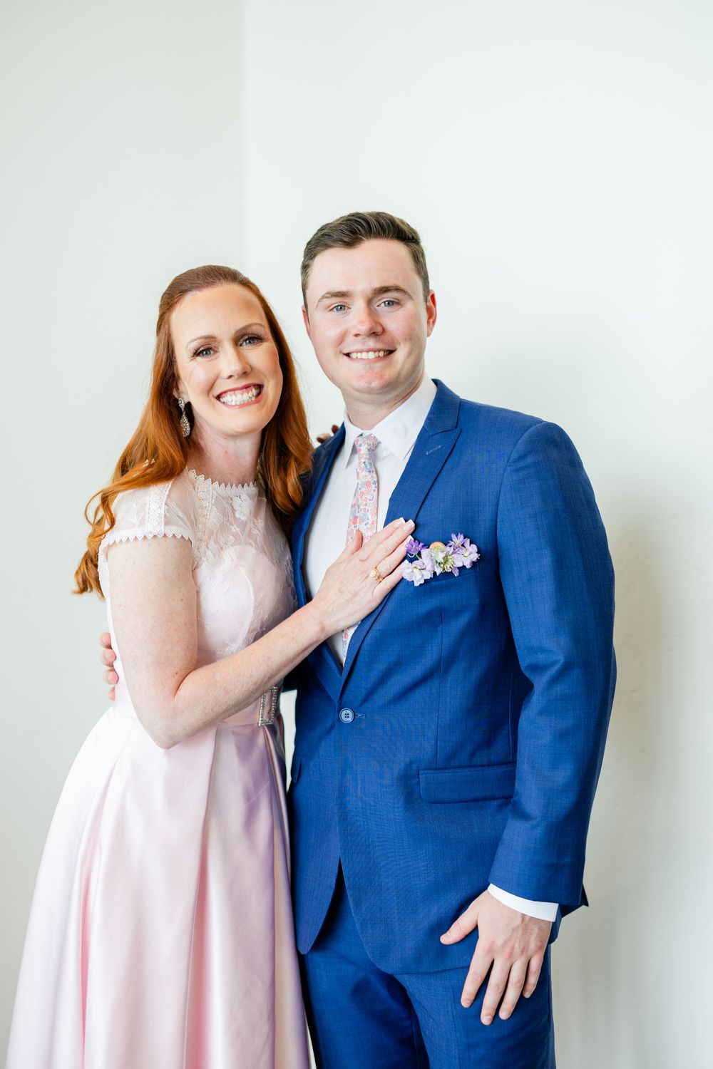 A couple poses together, with her in a pink dress and him in a royal blue suit with floral boutonniere.